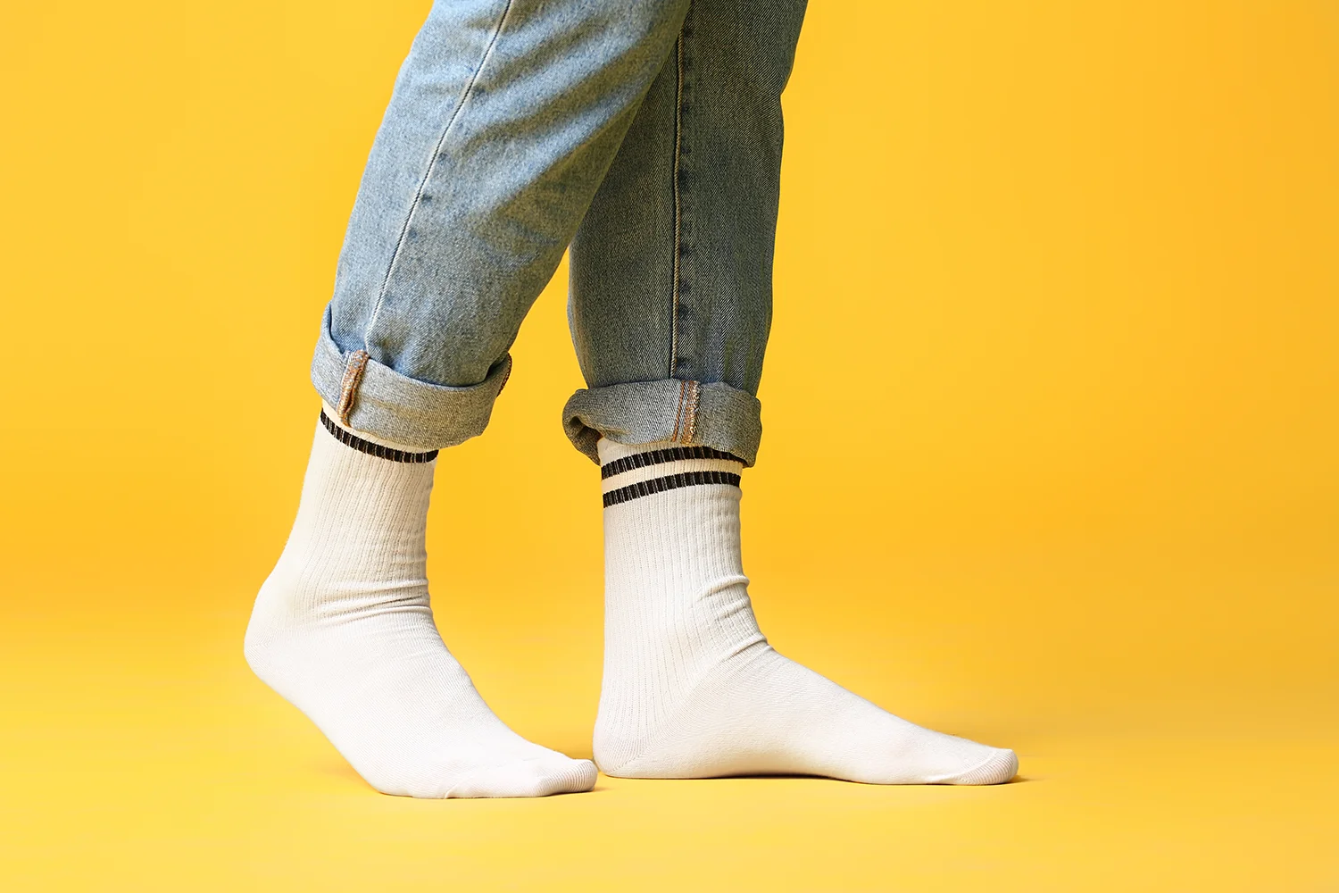Männliche Beine in hochgekrempelten Jeans und weißen Socken mit schwarzen Streifen vor gelbem Hintergrund