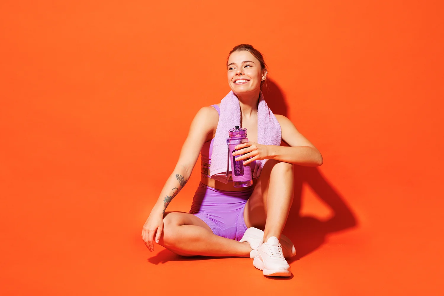 Frau in lila Sportoutfit sitzt vor orangefarbener Wand, hält lila Trinkflasche, lila Handtuch um den Hals