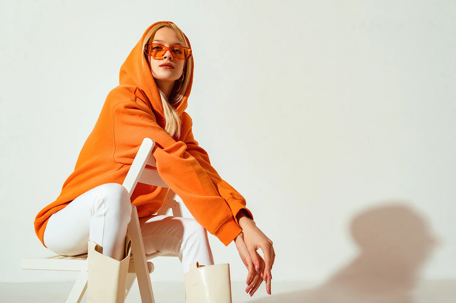 Frau sitzt auf weißem Stuhl, trägt orangefarbenen Hoodie, weiße Hose und cremefarbene Stiefel, orangefarbene Sonnenbrille