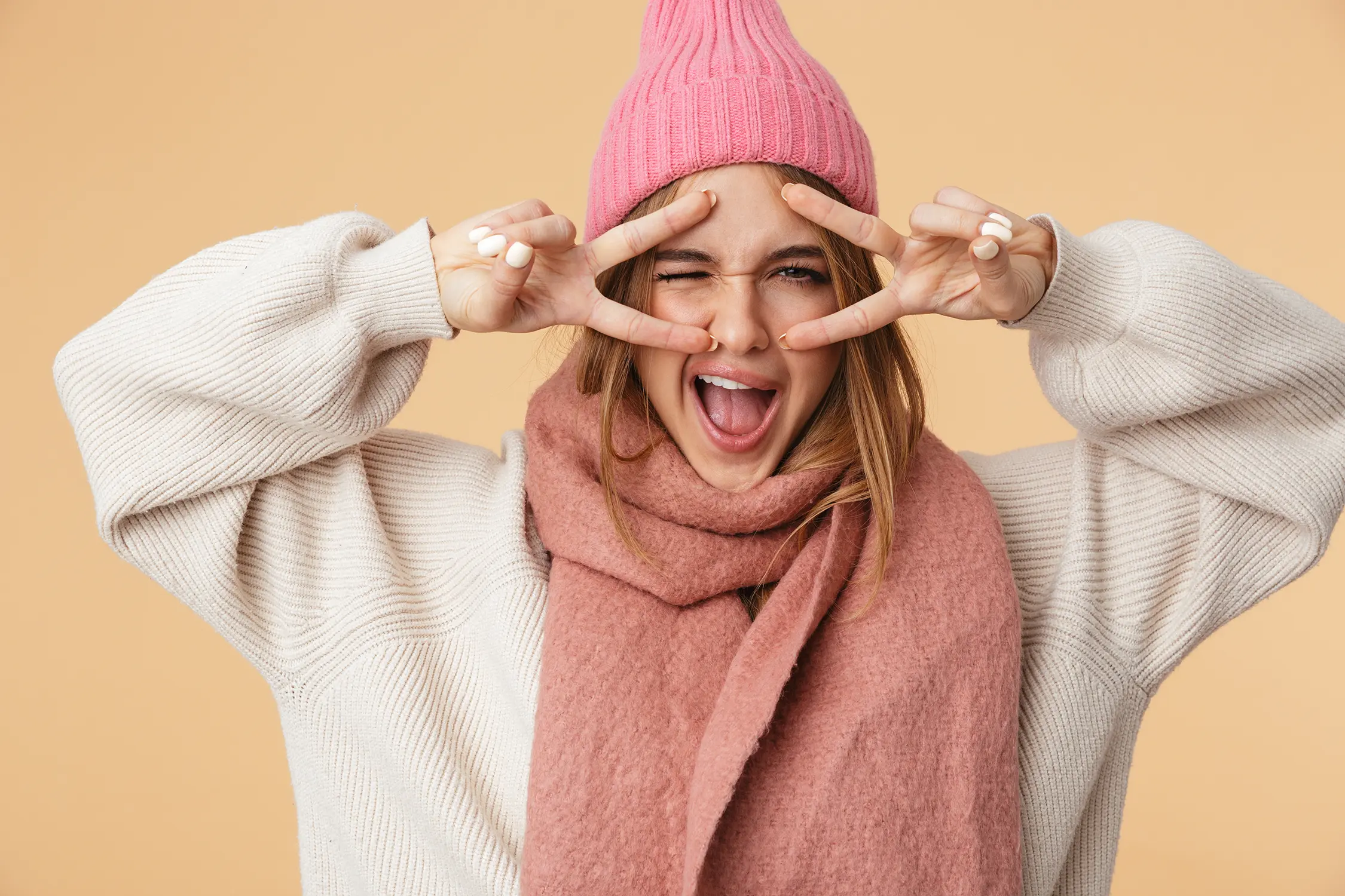 Frau in rosa Strickmütze, rosa Schal und cremefarbenem Pullover macht mit den Fingern ein Peace-Zeichen vor den Augen und lacht