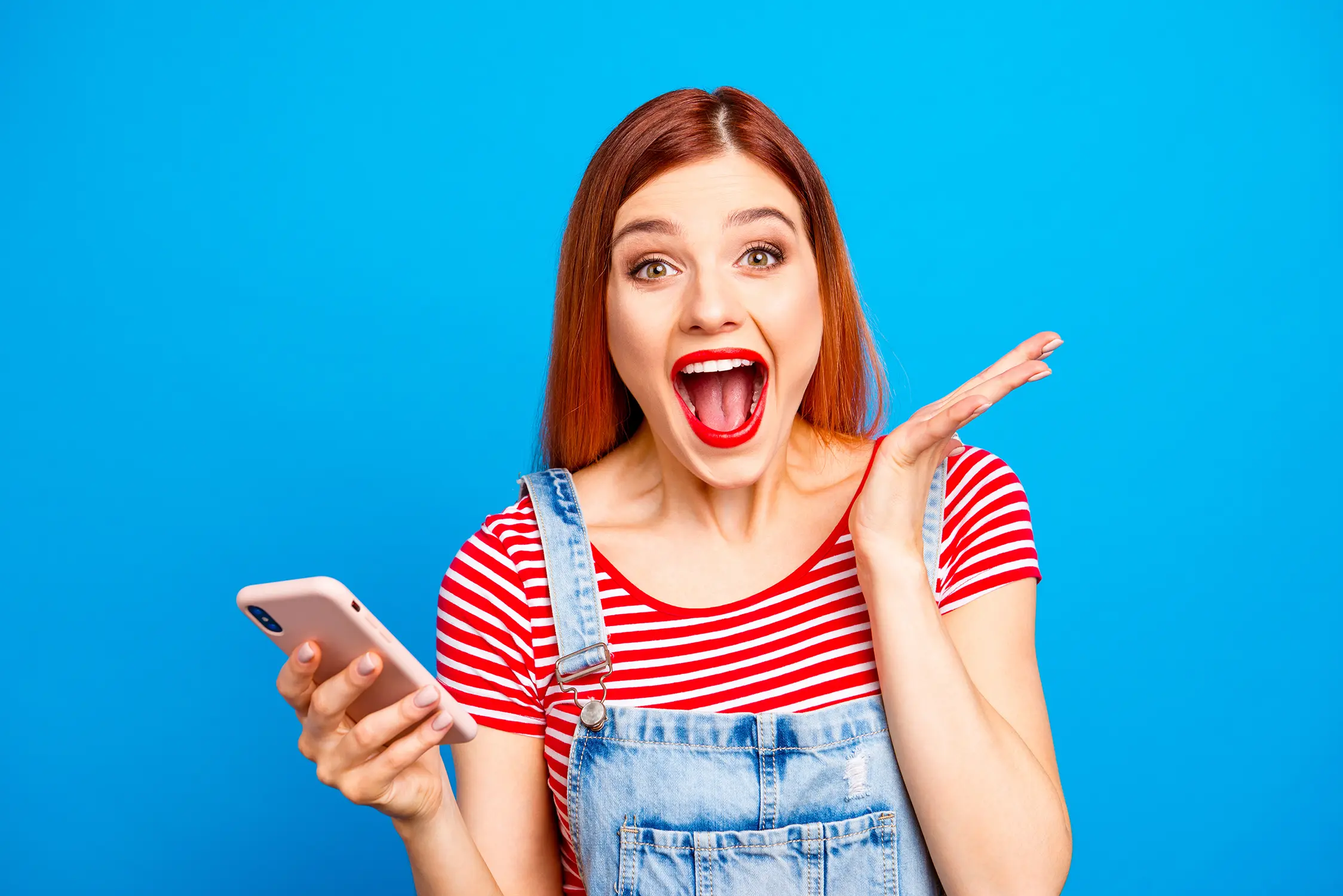 Junge Frau mit roten Lippen und gestreiftem Shirt hält Smartphone und zeigt überraschten Gesichtsausdruck vor blauem Hintergrund