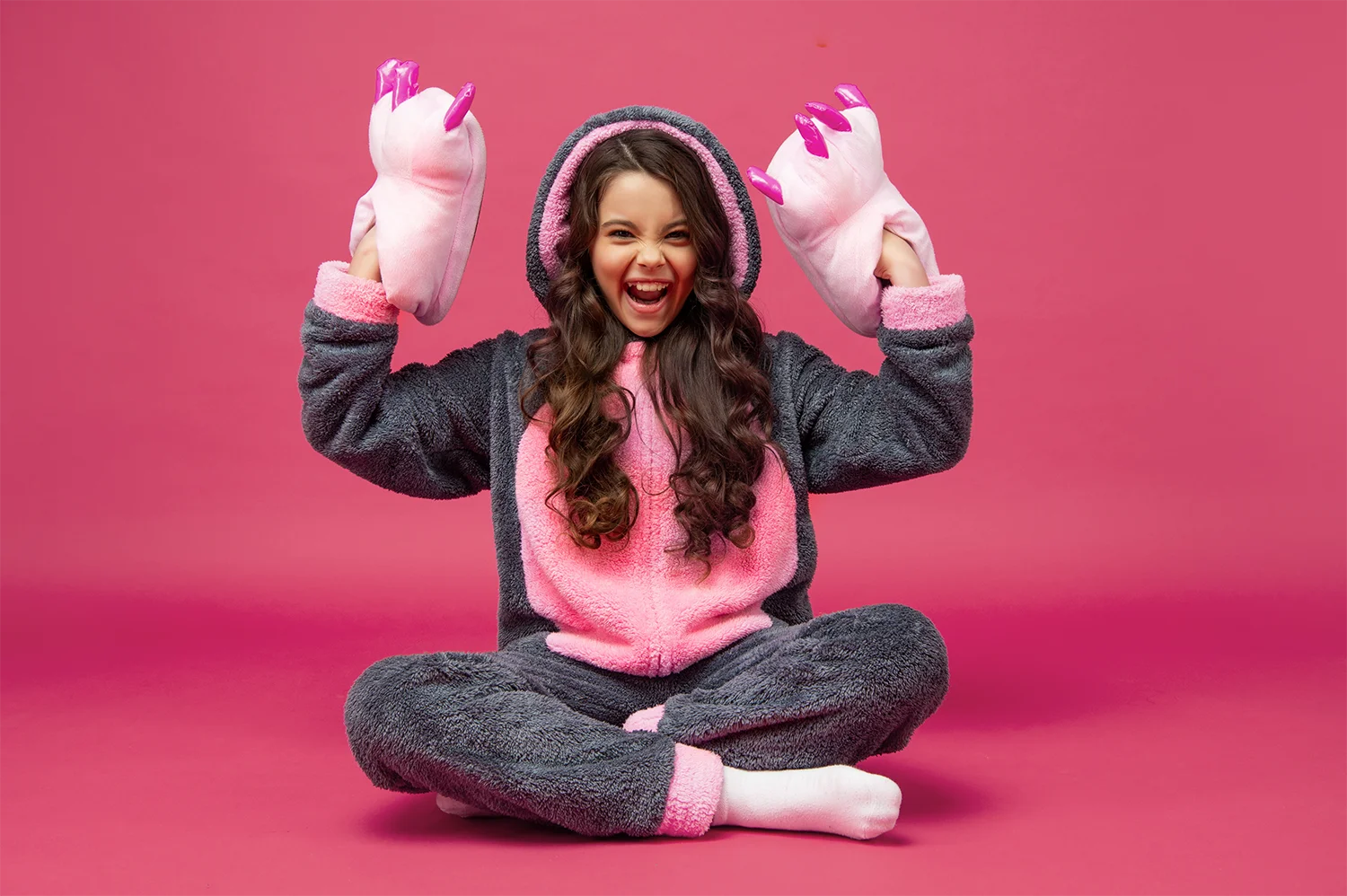 Mädchen in grauer und pinker Kuscheljacke mit Kapuze sitzt im Schneidersitz und hält pinke Hausschuhe mit langen Krallen hoch