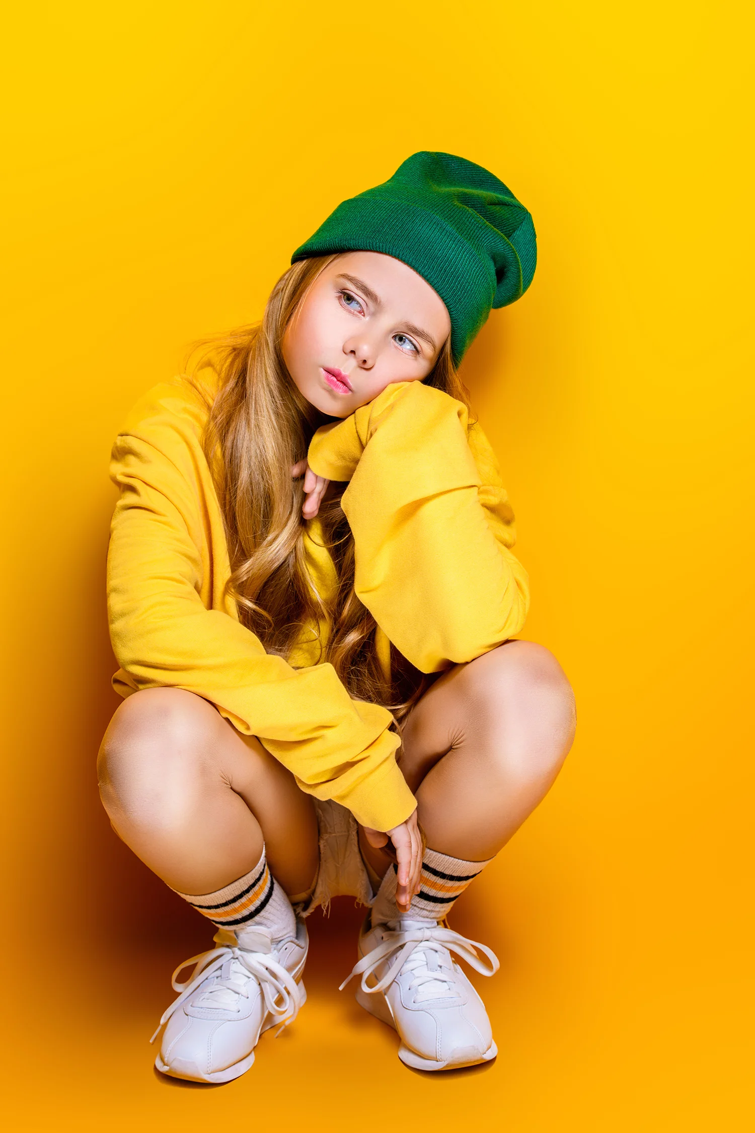 Mädchen mit grüner Mütze, gelbem Pullover, weißen Sneakers und gestreiften Socken sitzt in Hockstellung vor gelbem Hintergrund