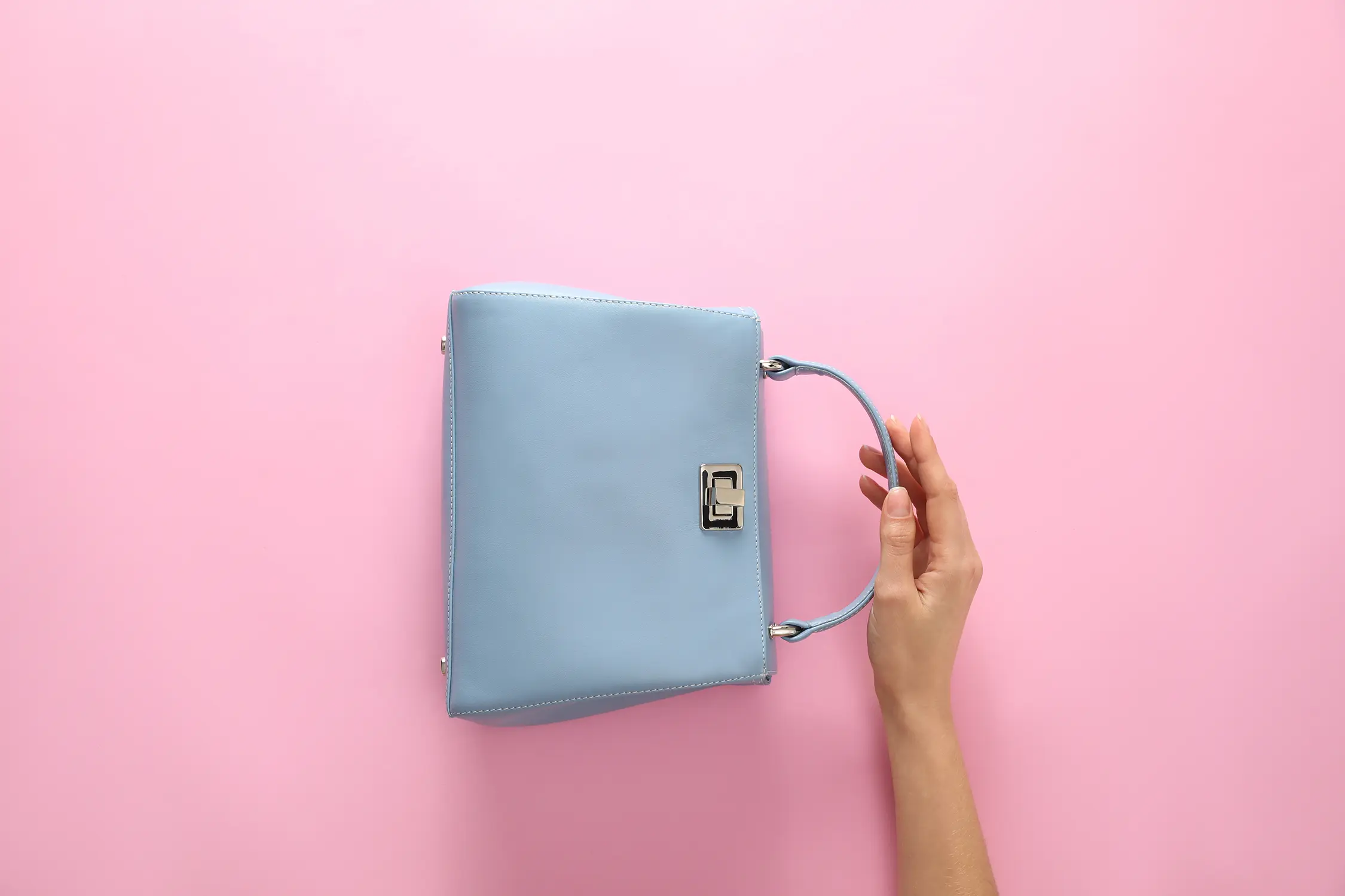 Hand hält hellblaue Handtasche mit silberner Schnalle vor rosa Hintergrund