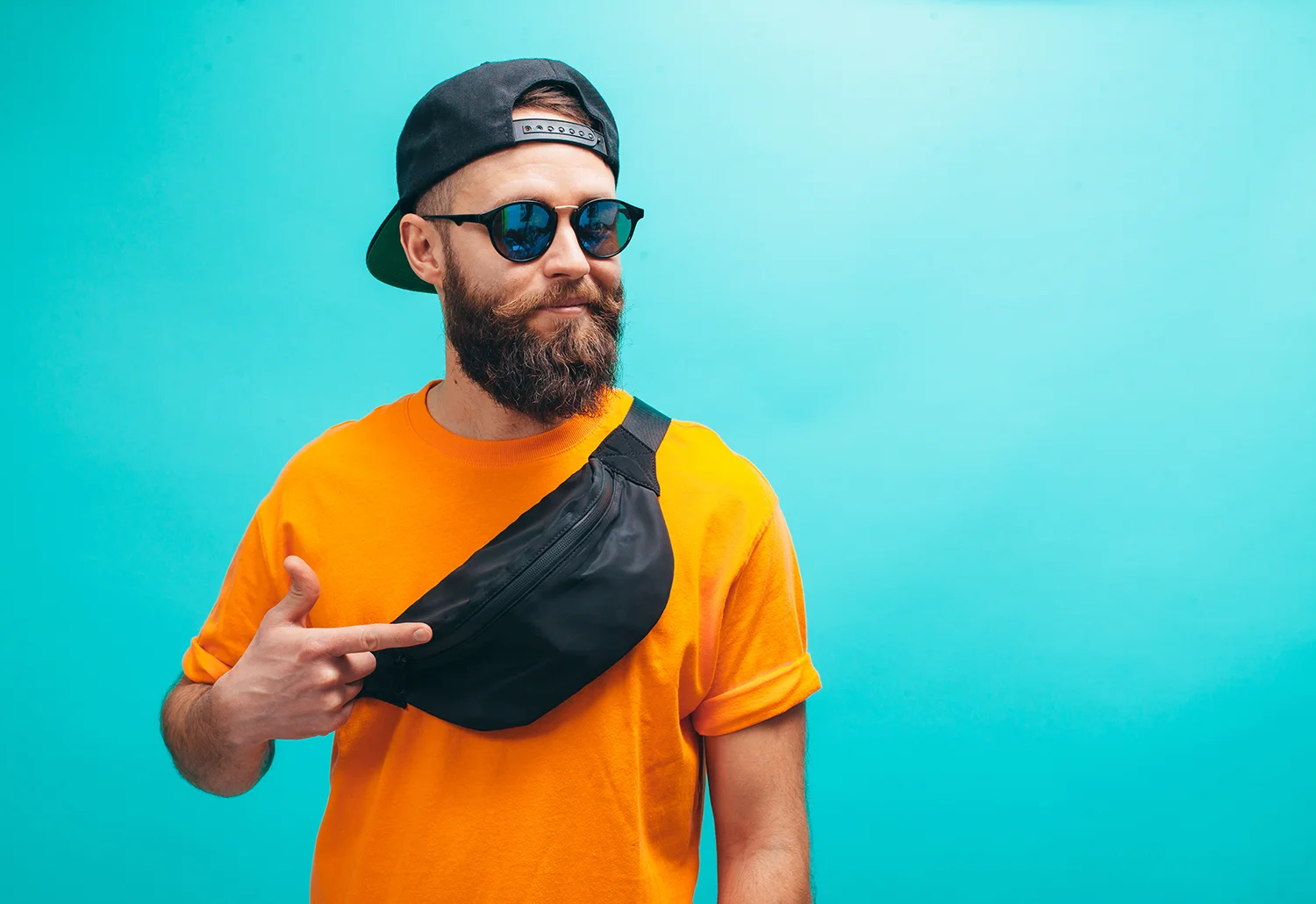 Mann mit Bart, schwarzer Kappe rückwärts, Sonnenbrille und orangefarbenem T-Shirt zeigt auf schwarze Bauchtasche vor blauem Hintergrund