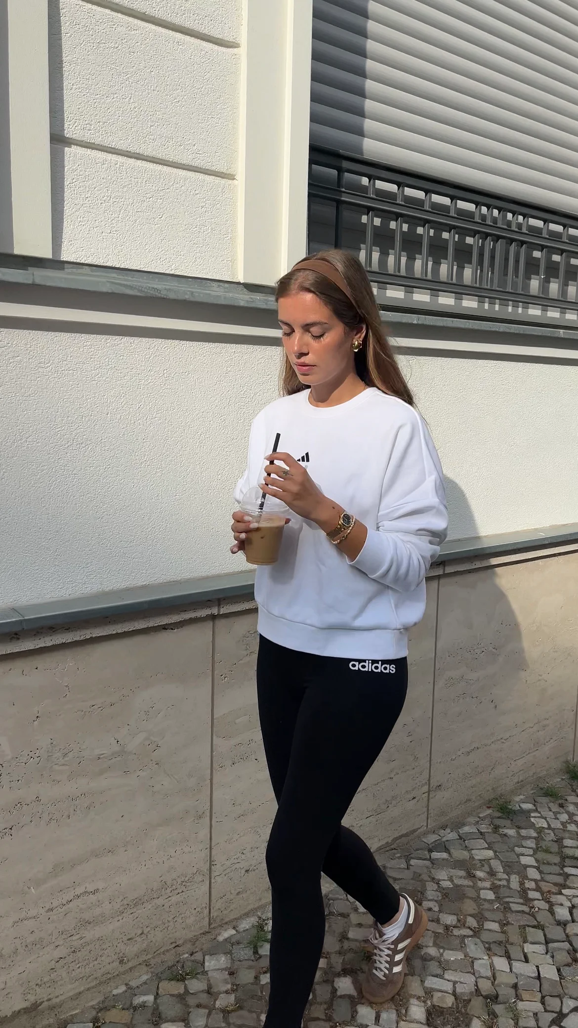 Frau in weißem Sweatshirt und schwarzer Adidas-Leggings geht mit Eiskaffee in der Hand an einer Hauswand entlang.
