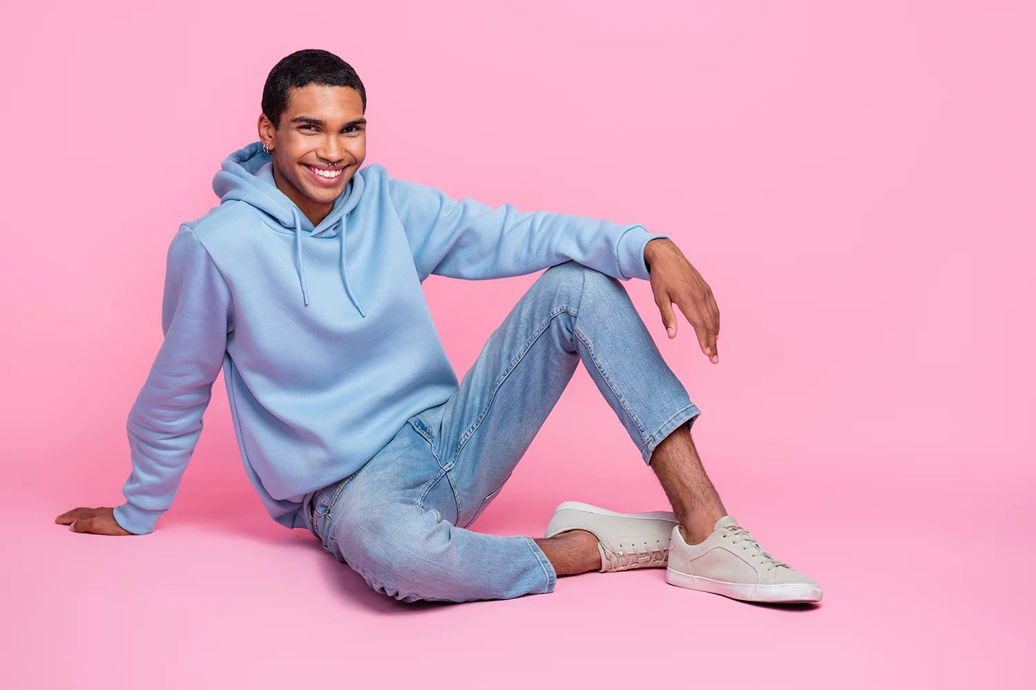 Junger Mann in hellblauem Hoodie, Jeans und weißen Casual-Schuhen sitzt lächelnd vor rosa Hintergrund