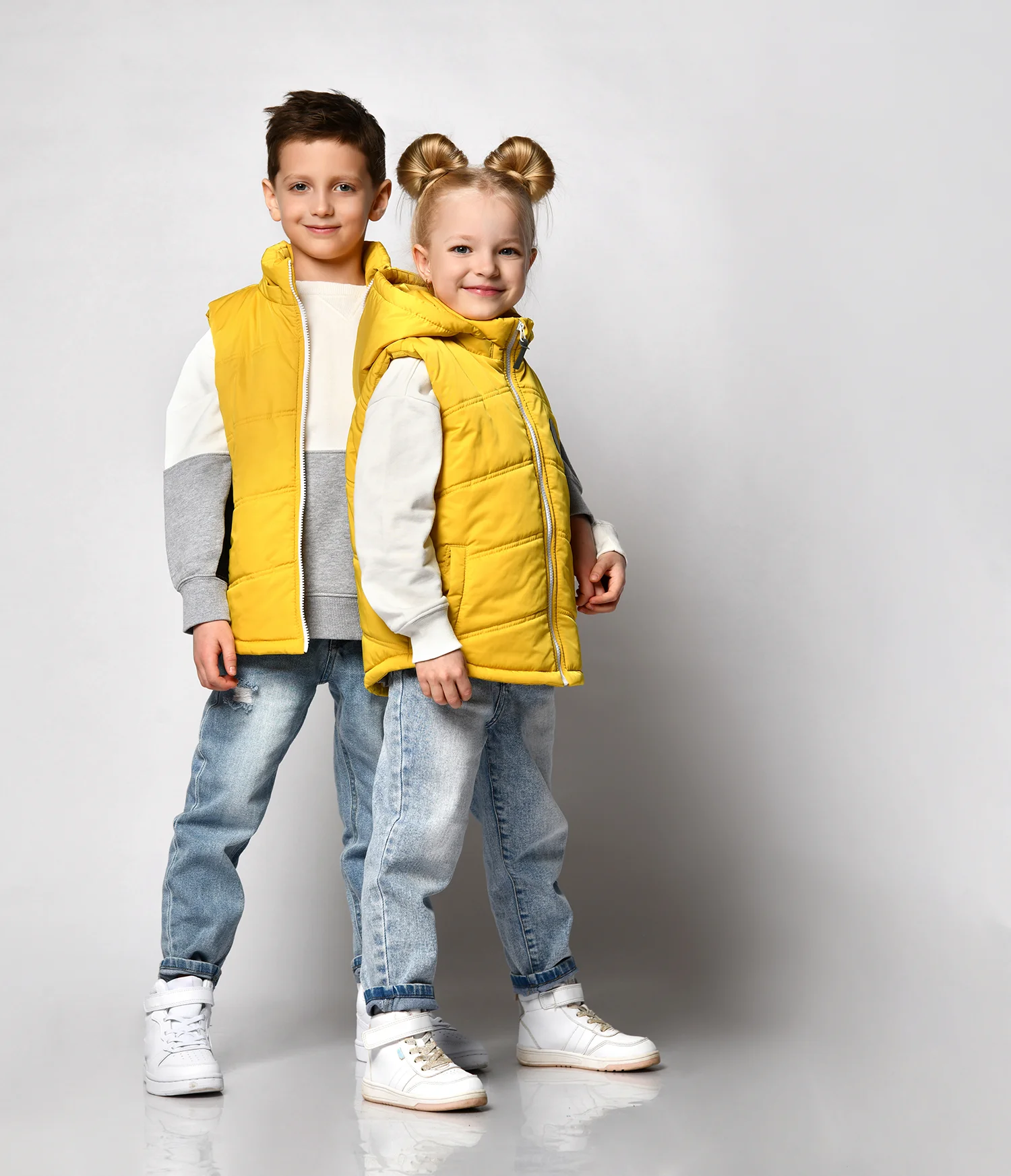 Zwei Kinder, Junge und Mädchen, tragen gelbe Westen, weiße Sweatshirts, blaue Jeans und weiße Sneaker vor grauem Hintergrund