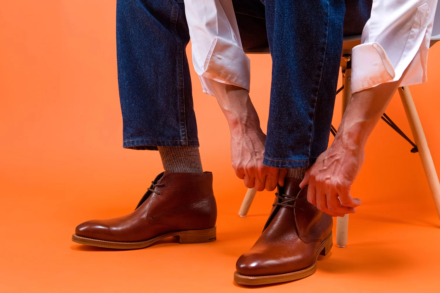 Mann bindet braune elegante Herrenschuhe aus Leder, trägt blaue Jeans und graue Socken, sitzt auf Stuhl vor orangefarbenem Hintergrund
