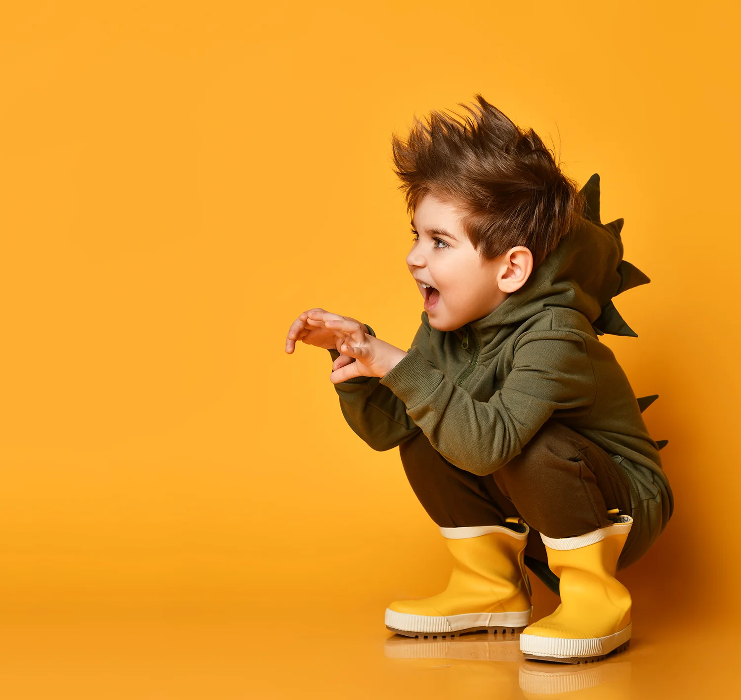Kleiner Junge in grüner Dino-Kapuzenjacke und gelben Gummistiefeln hockt und macht spielerische Klauenbewegung