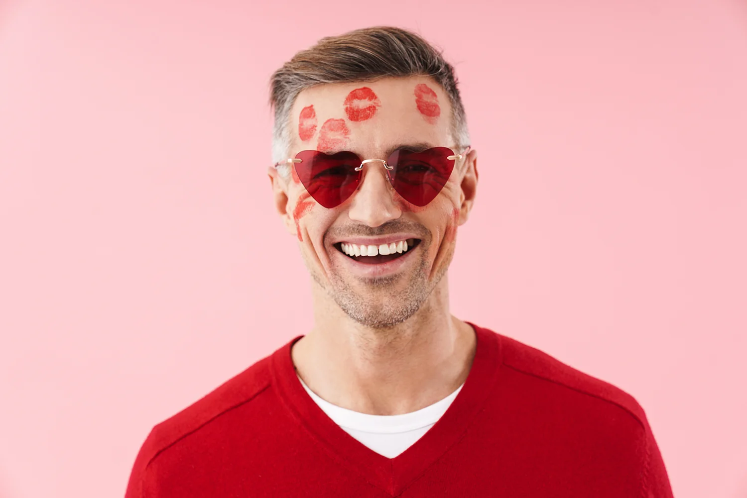 Lächender Mann mit roten herzförmigen Sonnenbrillen und roten Lippenabdrücken im Gesicht vor rosa Hintergrund