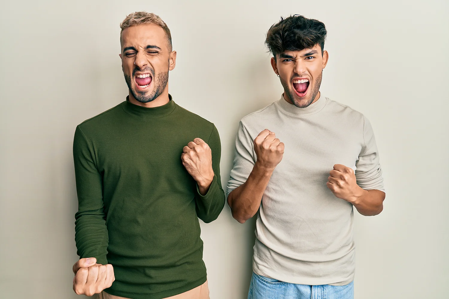 Zwei Männer mit geballten Fäusten zeigen Freude und Begeisterung, einer trägt grünen Pullover, der andere hellen Pullover und Jeans