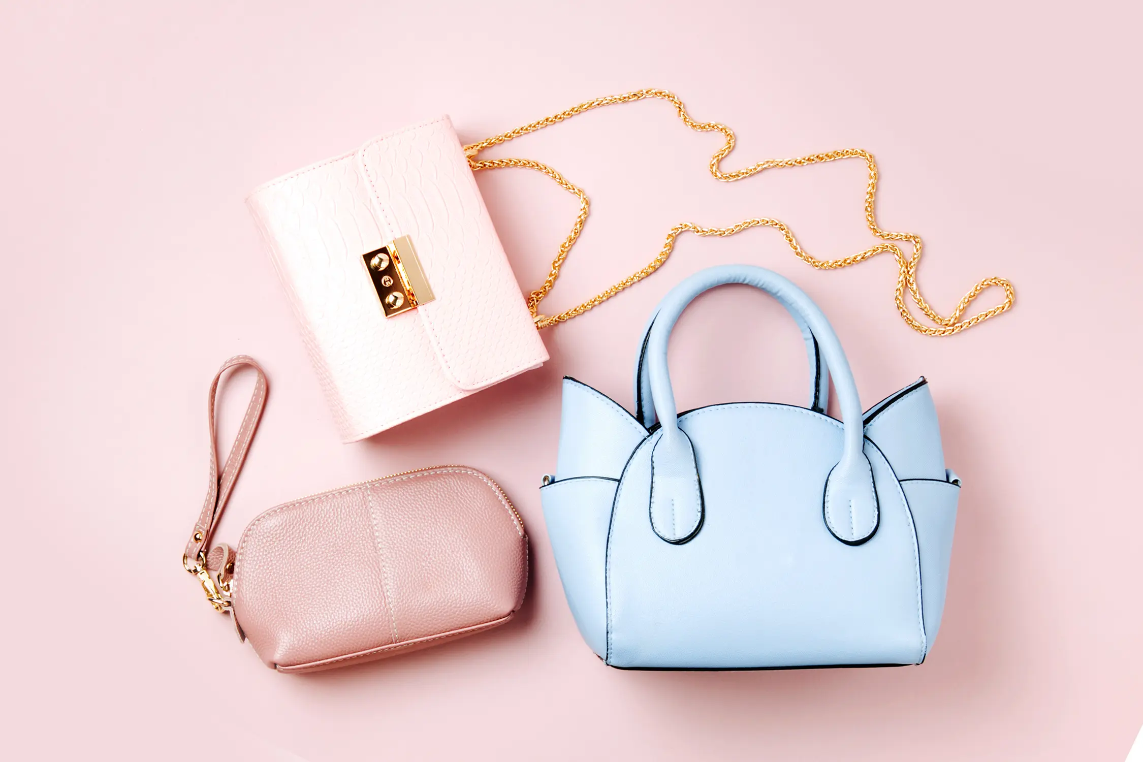 Drei modische Taschen und Accessoires auf rosa Hintergrund: eine kleine blaue Handtasche, eine rosa Clutch mit goldener Kette und ein kleines rosa Täschchen mit Reißverschluss