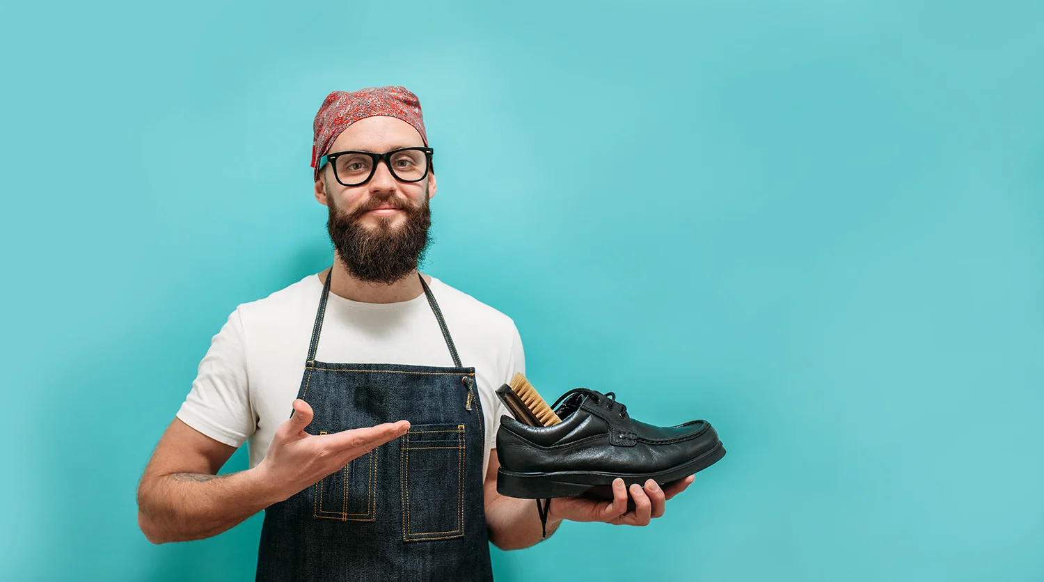 Mann mit Bart, Brille und rotem Kopftuch hält schwarzen Lederschuh und Schuhbürste vor blauem Hintergrund