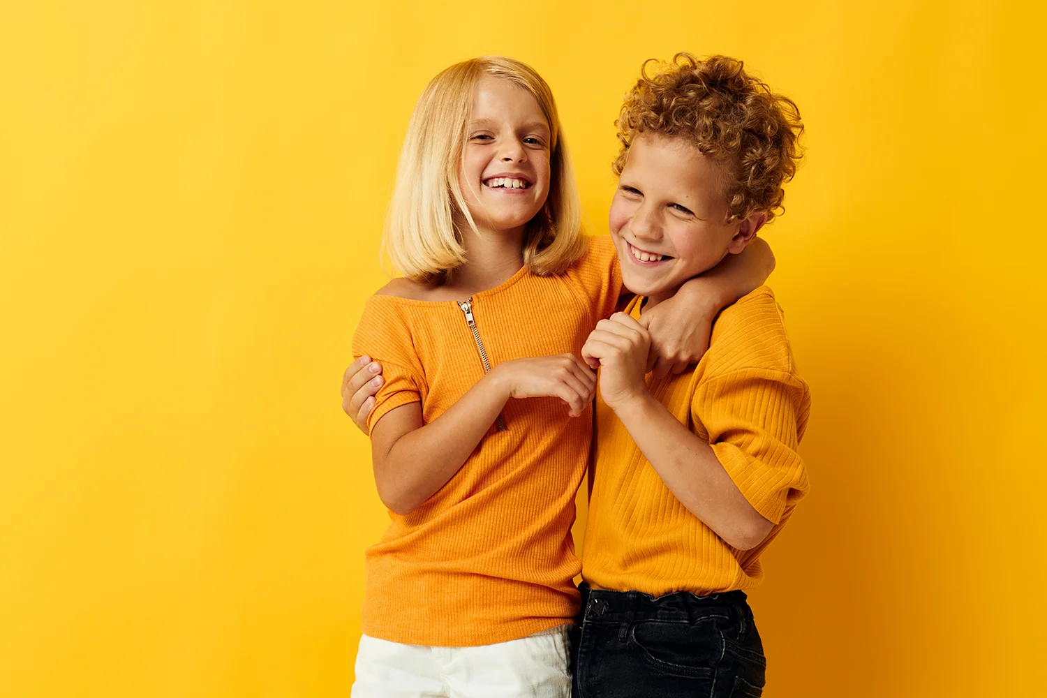 Zwei Kinder in orangefarbenen Oberteilen umarmen sich vor gelbem Hintergrund