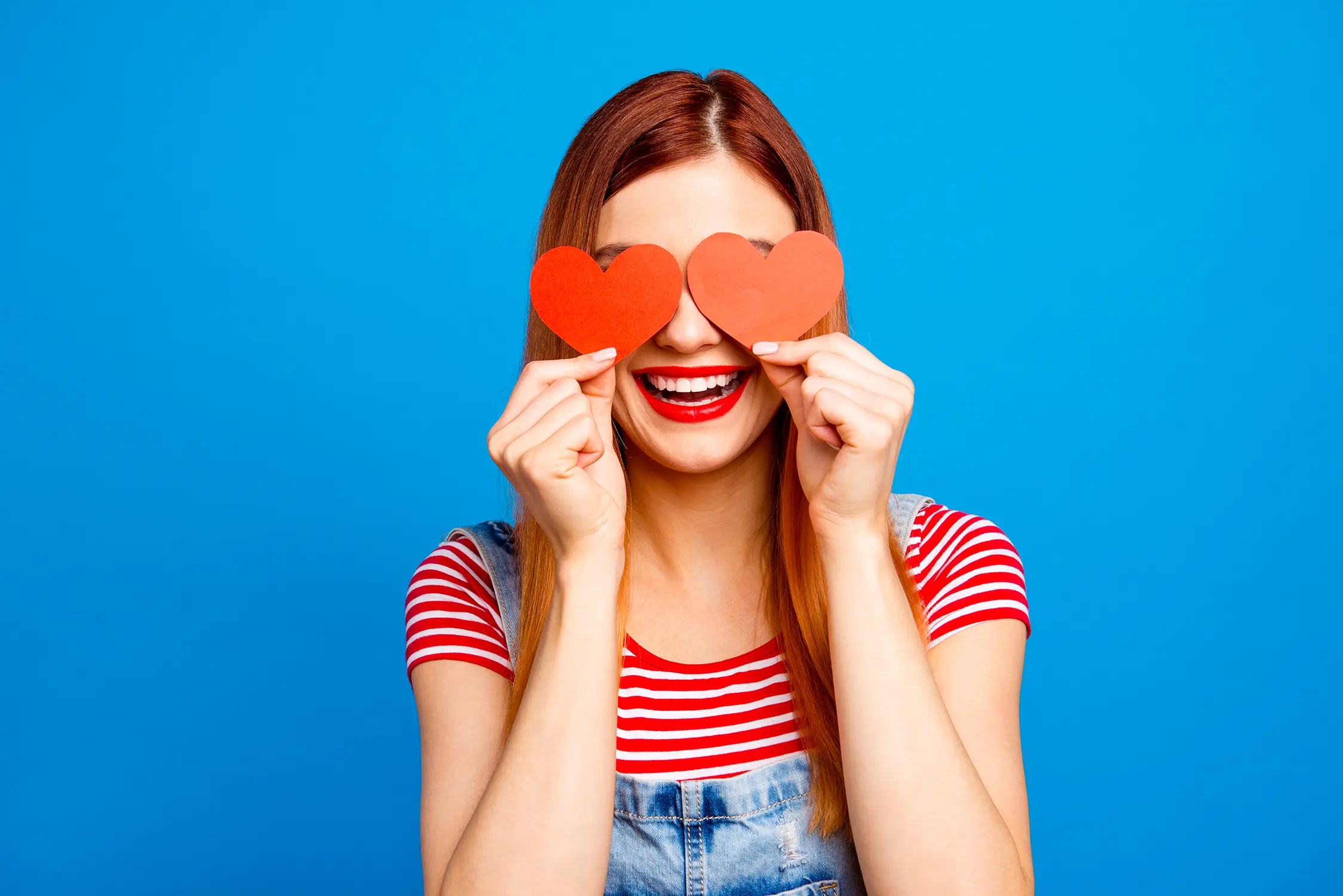 Frau mit roten Haaren und rotem Lippenstift hält zwei rote Papierherzen vor ihre Augen, trägt rot-weiß gestreiftes Shirt und Jeans-Latzhose vor blauem Hintergrund