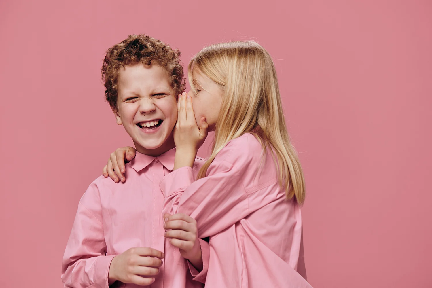 Mädchen flüstert Junge etwas ins Ohr, beide tragen rosa Hemden vor rosa Hintergrund