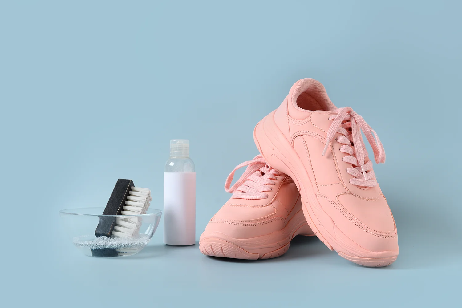 Rosa Sportschuhe neben einer kleinen Flasche und einer Bürste in einer Schüssel mit Seifenwasser auf blauem Hintergrund